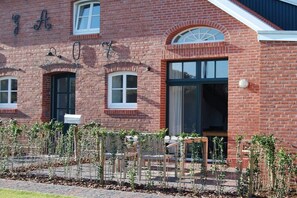 Een bakstenen gebouw met een groot raam en een deur, een brievenbus, en een kleine tuin met planten en een bankje.