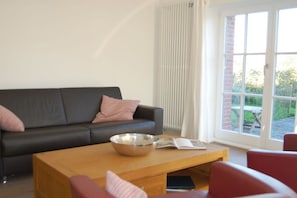 A modern living room with a dark sofa, a wooden coffee table, and a view through a large window. - Holiday apartment with 2 bedrooms (Langeoog)