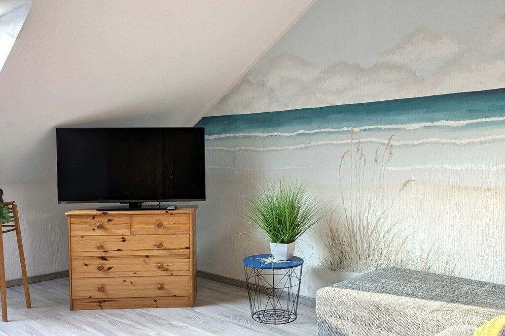 A room with a television on a wooden dresser, a small round table with a plant, and a wall mural of a beach scene.