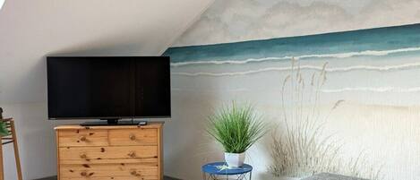 A room with a television on a wooden dresser, a small round table with a plant, and a wall mural of a beach scene.