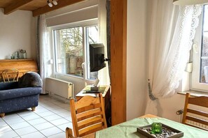 A room with a wooden ceiling, a blue sofa, a wooden table with a green cloth, and a window with curtains. - Holiday home with 3 bedrooms (Wangerland-Hooksiel)