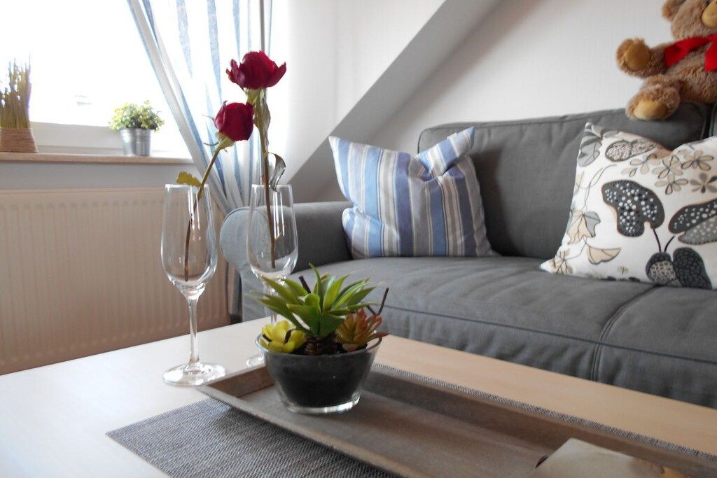 Un salon avec un canapé gris, des coussins à rayures, un ours en peluche, un vase rempli de roses rouges et une petite plante posée sur une table.