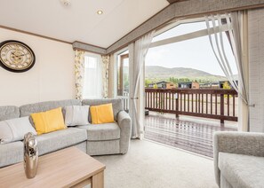 A living room with a grey sofa, yellow cushions, a wooden coffee table, and a large window with a view of a balcony and buildings. - Modern two-bedroom holiday home. (Fort Augustus)