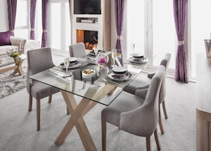 A modern dining area with a glass table, grey upholstered chairs, and a fireplace in the background. - 1 bedroom accommodation in Fort Augustus (Fort Augustus)