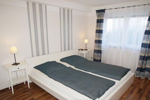 A double bed with a white headboard and a grey mattress, a bedside table with a lamp, and a window with sheer curtains. - Holiday apartment with 1 bedroom (Fehmarn)