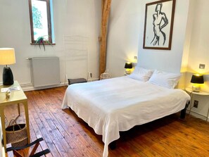 A bedroom with a large bed, wooden flooring, a radiator, a window with a view, and a painting on the wall.
