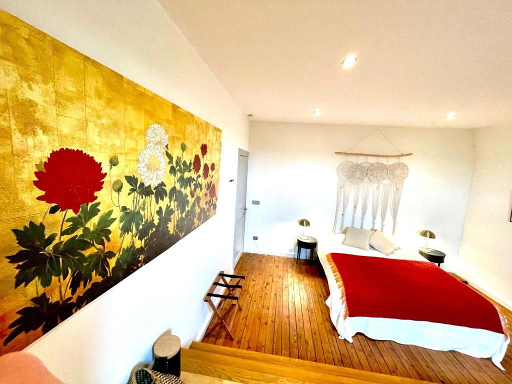 A room with a bed, a small table, a chair, a wall with a floral painting, and a hanging light fixture.