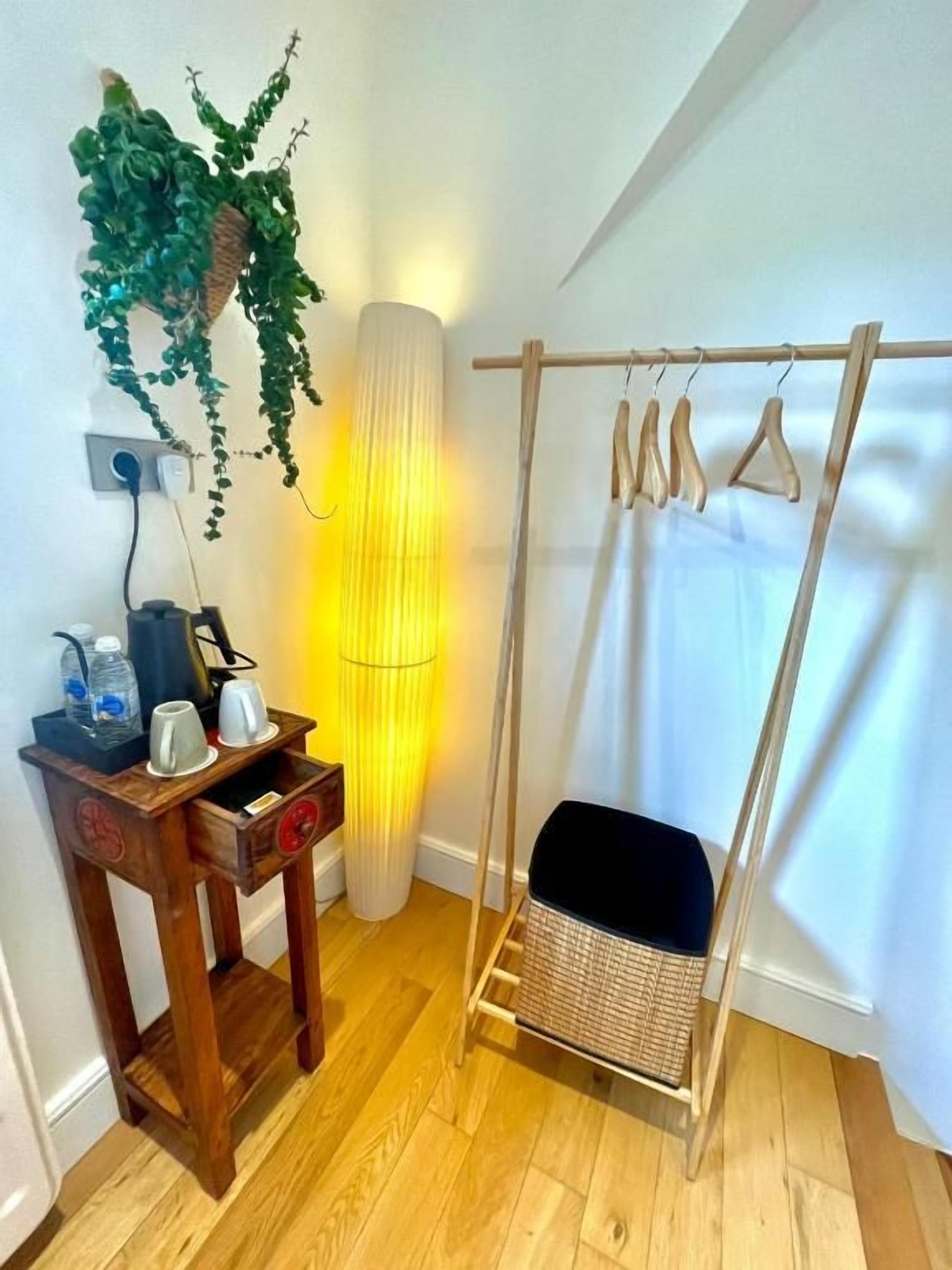 A room with a wooden floor, a hanging plant, a lamp, a wooden table with items, and a swing with a basket seat.