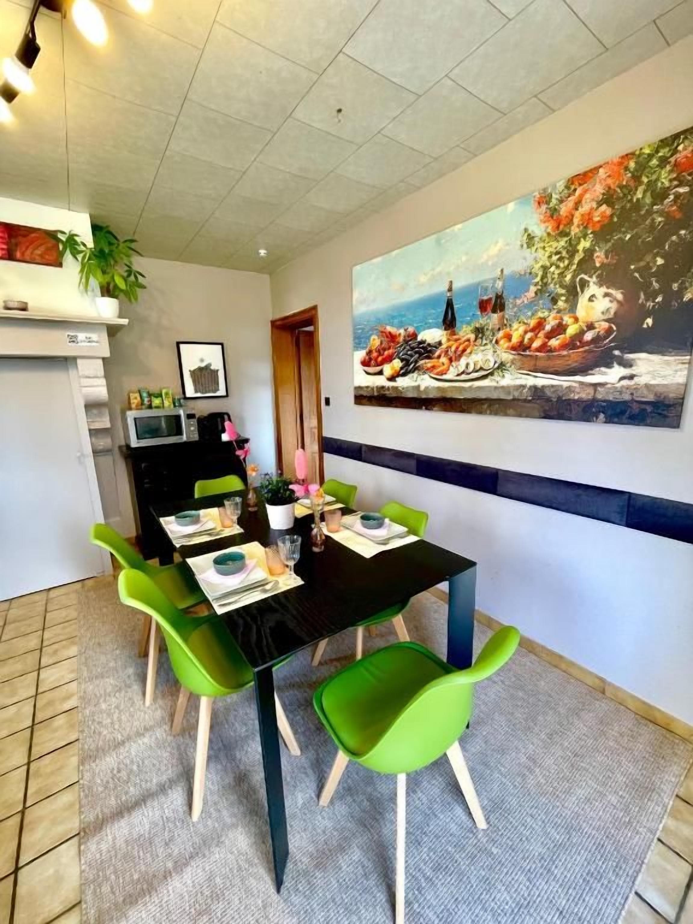 Un coin repas avec une table noire, des chaises vertes et un tableau représentant des fruits et des fleurs accroché au mur.