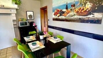 Un coin repas avec une table noire, des chaises vertes et un tableau représentant des fruits et des fleurs accroché au mur.