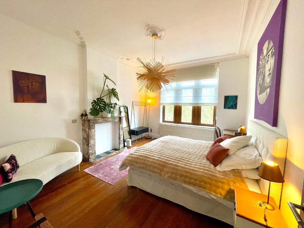 Un dormitorio con cama, una silla, ventana con cortinas, una planta y una obra de arte colgada en la pared.