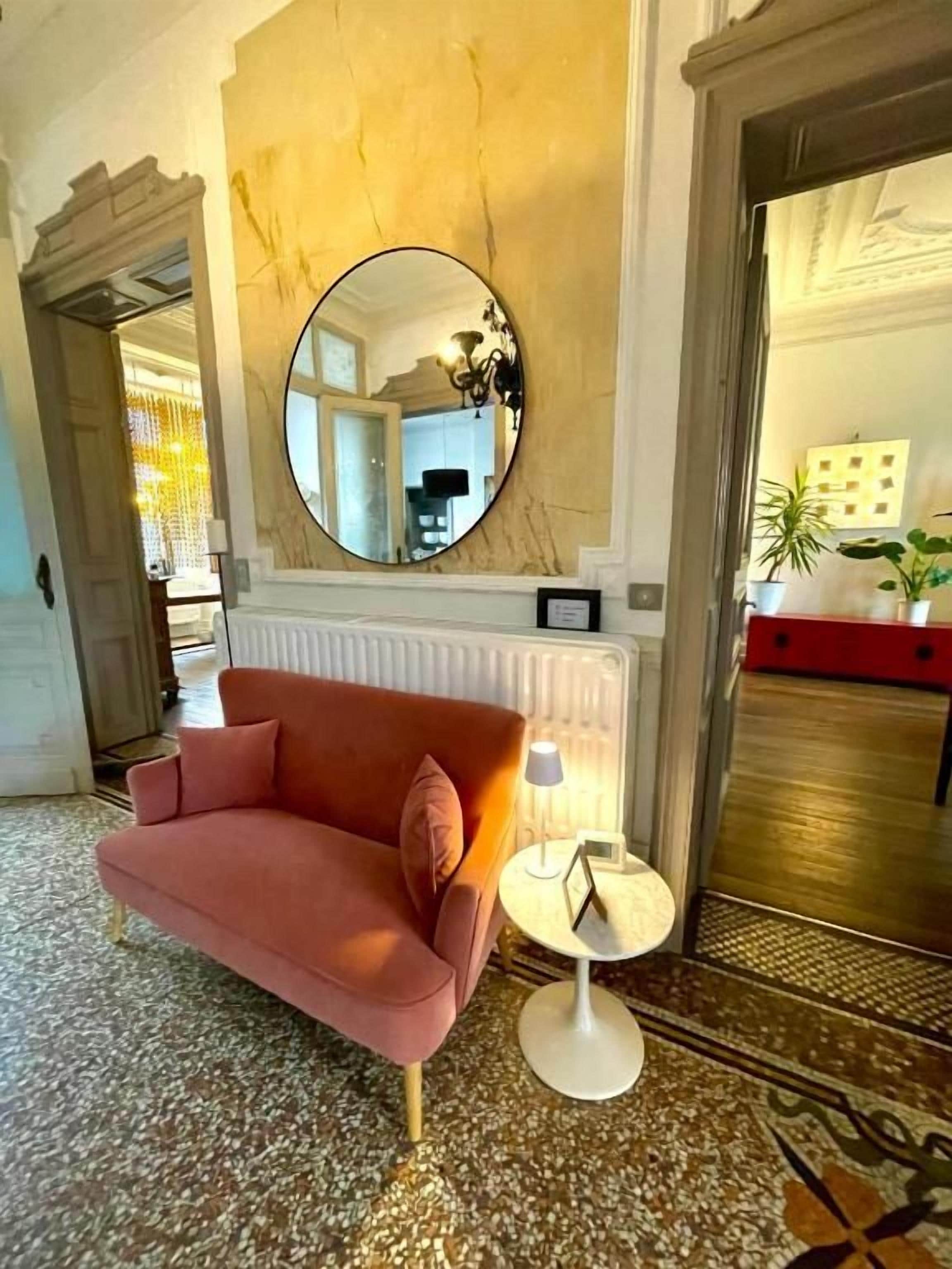 Un hall d’entrée avec un canapé rouge, un miroir rond, une petite table avec une lampe et un radiateur.