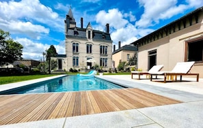 A swimming pool with a wooden deck, lounge chairs, and a large building with a tower in the background. - Villa O (Virton)