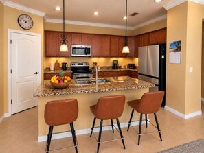 A kitchen with wooden cabinets, stainless steel appliances, a fruit bowl, and a bar area with stools. - Furnished Pool Home with Beautiful Lake Views in Stoneybrook at Venice (Venice)
