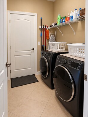 A laundry room with two front-loading washers and dryers, a shelf with cleaning supplies, and a door with a doormat. - Furnished Pool Home with Beautiful Lake Views in Stoneybrook at Venice (Venice)