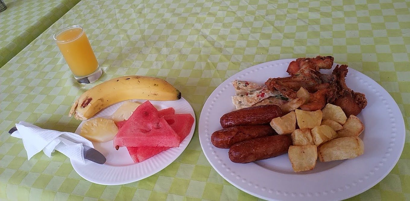 A table with a checkered tablecloth, a plate with a banana, watermelon, and a sausage dish, and a glass of orange juice.