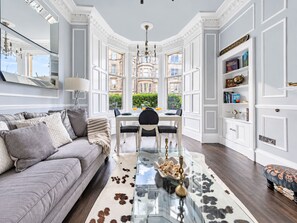 A room with a sofa, a dining table, and a view of buildings through large windows. - Lovely Flat near Royal Botanic Garden (Edinburgh)