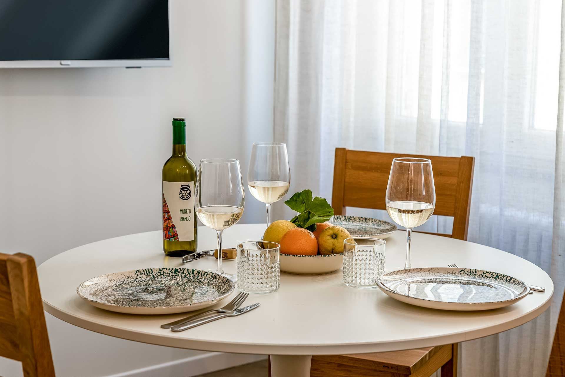 Un tavolo da pranzo apparecchiato con piatti, posate, una bottiglia di vino, bicchieri e una ciotola di frutta.