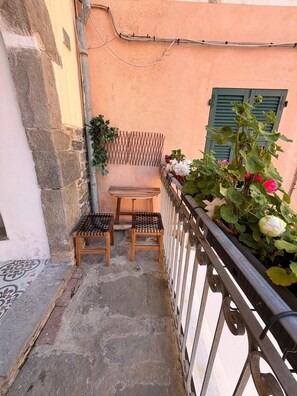Een klein buiten zitgedeelte met twee houten krukken en een bank, een metalen hekwerk, en een bloeiende plant in een pot.
