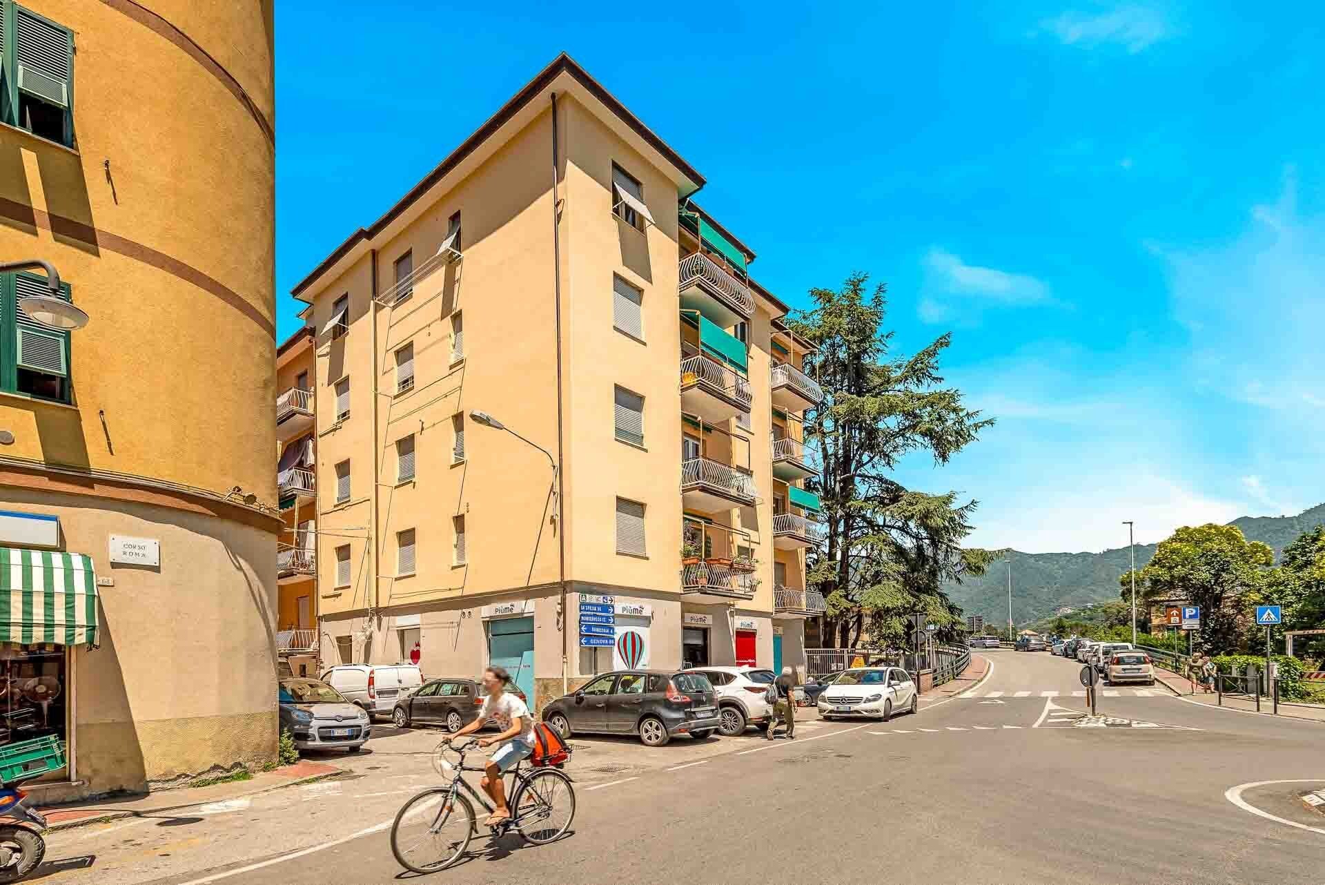 Ein mehrstöckiges Gebäude mit Balkonen, eine Straße mit Autos und einem Radfahrer, und ein klarer, blauer Himmel.
