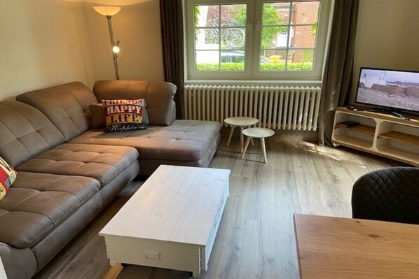 Ein Wohnzimmer mit einem braunen Ledersofa, einem weißen Couchtisch, einem kleinen weißen Beistelltisch, einem Fernseher auf einem Holzboden und einem Fenster mit Vorhängen.
