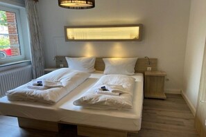 A double bed with white linens, a bedside table, a wall-mounted light, and a window with curtains. - Holiday apartment with 1 bedroom (Wyk auf Föhr)