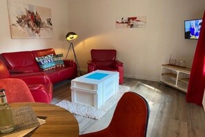 Ein Wohnzimmer mit einem roten Sofa, einem weißen Couchtisch, einem roten Sessel, einer Stehlampe und einem Fernseher an der Wand.