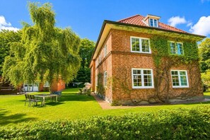 A two-story brick house with a red-tiled roof, surrounded by a well-maintained lawn and a large tree. - Holiday apartment with 1 bedroom (Wyk auf Föhr)