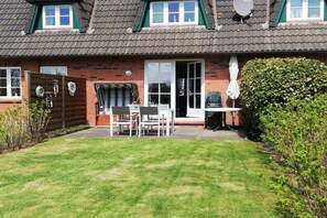 Ein Haus mit Ziegelaußenwand, einem Ziegeldach und einer Haustür. Vor dem Haus befindet sich eine kleine Terrasse mit Tisch und Stühlen, und eine Hecke auf der rechten Seite.
