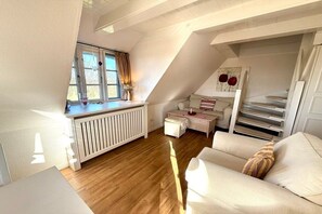 A cozy attic bedroom with a sloped ceiling, a white radiator, a window with curtains, a sofa, and a small table. - Deichhof Holiday Apartment 8 (Dunsum)