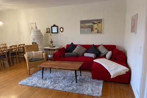 A living room with a red sofa, a wooden coffee table, a white rug, and a dining area with chairs and a table. - Holiday apartment with 2 bedrooms (Midlum auf Föhr)