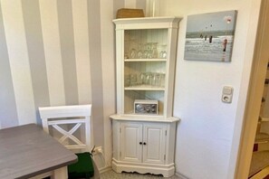 Ein Eckschrank mit Gläsern und einem Radio, ein weißer Stuhl und ein Gemälde mit einer Strandansicht.