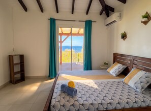 A bedroom with a bed, a wooden headboard, a nightstand, a potted plant, and a view of the sea through a glass door. - Homerez - House Baillif (Baillif)