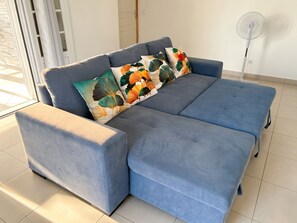 A blue sofa with a floral cushion, a standing fan, and a window with blinds. - Homerez - House Baillif (Baillif)