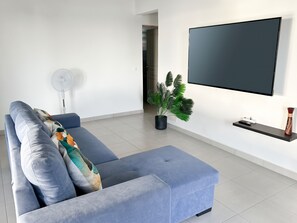 A modern living room with a blue sofa, a flat-screen TV mounted on the wall, a standing fan, and a potted plant. - Homerez - House Baillif (Baillif)
