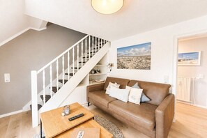 A living room with a brown leather sofa, a wooden coffee table, a white staircase, and a framed landscape painting on the wall. - Holiday apartment Ankerplatz Wrixum (Wrixum auf Föhr)