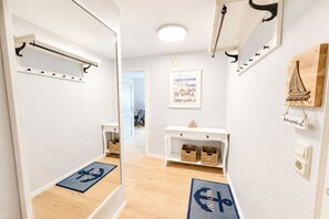 A hotel hallway with a coat rack, a framed picture, a small table with baskets, and a doormat with a nautical symbol. - Holiday apartment Ankerplatz Wrixum (Wrixum auf Föhr)