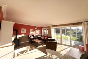 A living room with a fireplace, a dining area, and a large sliding glass door leading to a garden. - Holiday home in Wyk with swimming pool (Wyk)