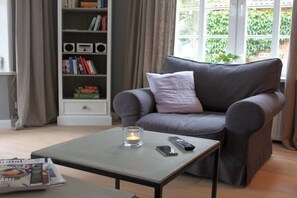 A living room with a grey sofa, a coffee table, a bookshelf, and a window with greenery outside. - Holiday home in Föhr with 1 bedroom (Wyk auf Föhr)