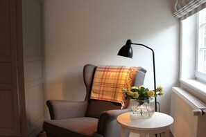 A room with a single armchair, a small table with a vase of flowers, a black floor lamp, and a window with curtains. - Holiday home in Föhr with 1 bedroom (Wyk auf Föhr)