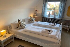 Cama doble con ropa de cama blanca y almohadas decorativas, una mesita de noche con lámpara y una ventana con cortinas.