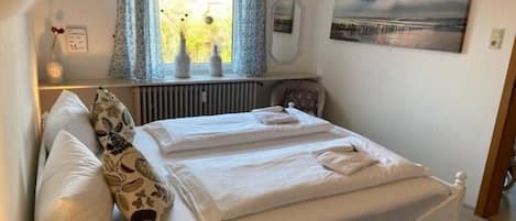 A neatly made double bed with white linens, a bedside table with a vase, and a window with curtains.