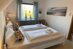 A neatly made double bed with white linens, a bedside table with a vase, and a window with curtains. - Holiday apartment in Föhr with 2 bedrooms (Wyk auf Föhr)
