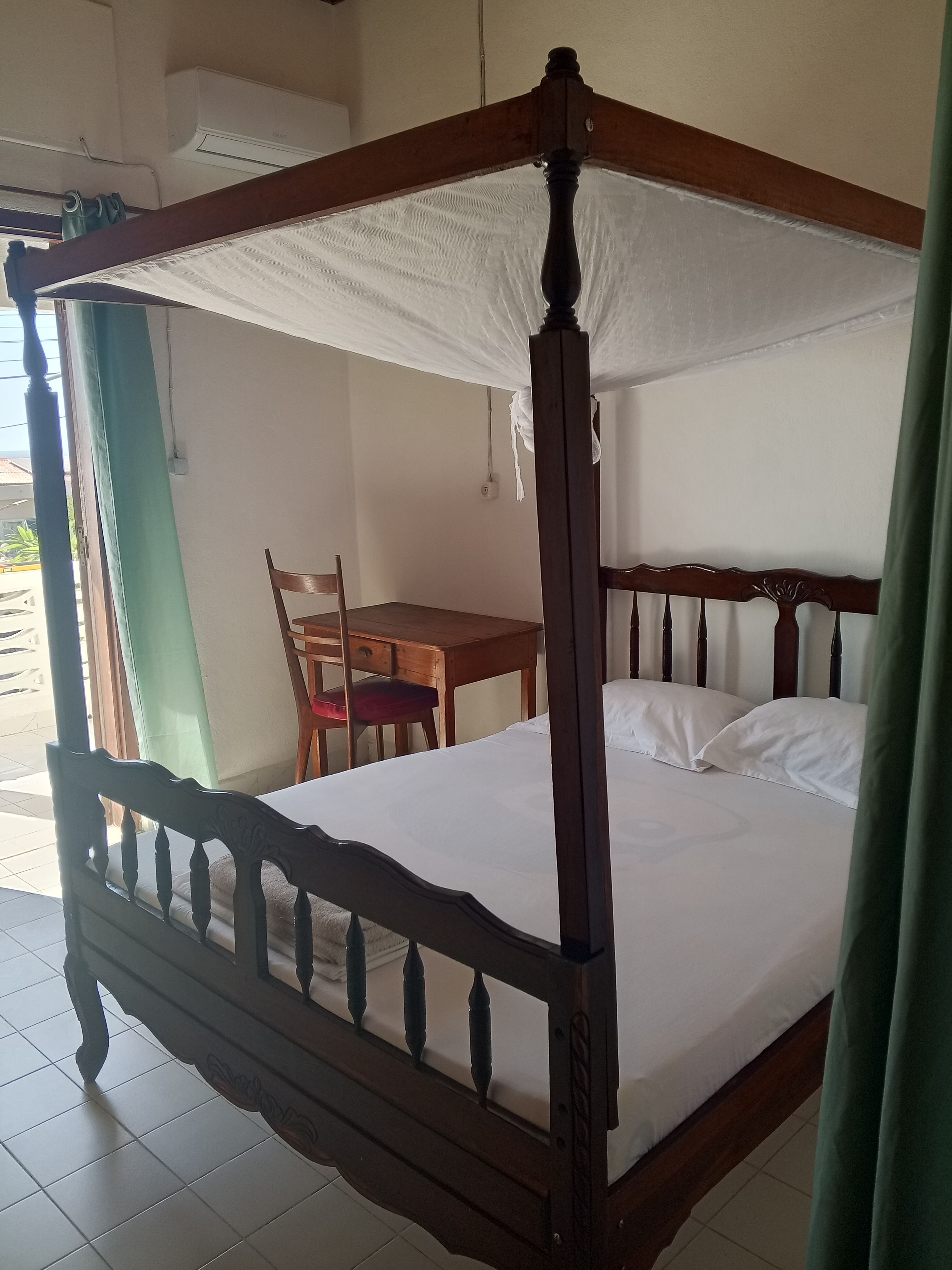 A four-poster bed with a canopy, a wooden table, and a chair in a room with tiled flooring.