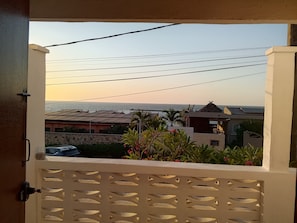 Blick von einem Balkon mit weißem Gitterzaun auf eine Küstenlandschaft mit Gebäuden und blauem Himmel.