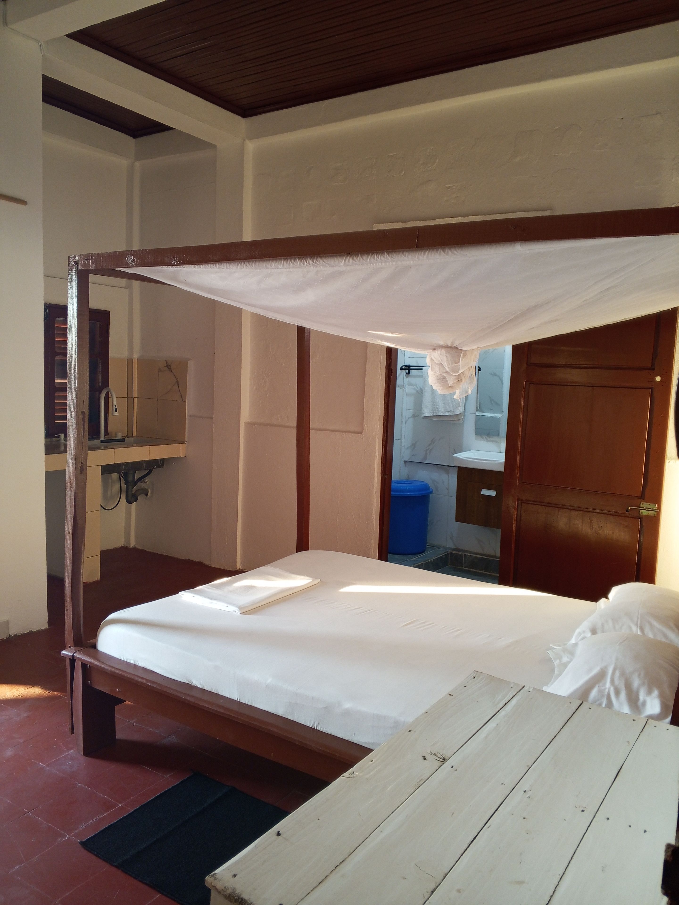 A single-bed room with a wooden frame, a canopy bed, a bedside table, and a view of a bathroom with a sink and mirror.