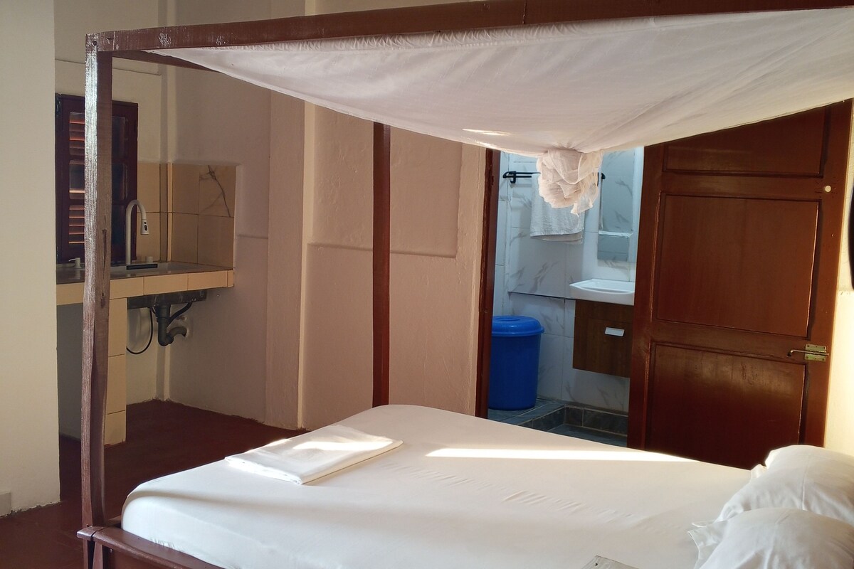 Einzimmer mit Einzelbett, Holzrahmen, Baldachin, Nachttisch und Blick auf ein Badezimmer mit Waschbecken und Spiegel.