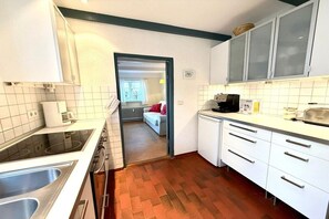 Eine Küche mit weißen Schränken, einem Herd und einem Spülbecken. Ein Durchgang führt in ein Schlafzimmer mit einem Bett und einem Fenster.