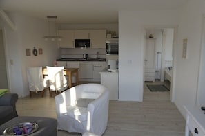 A modern living room with a white sofa, a dining table, and a kitchen area with white cabinets and appliances. - Holiday apartment in Föhr with 1 bedroom (Oldsum)