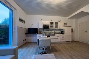 A modern kitchen with white cabinetry, a dining area with a table and chairs, and a large window providing a view of the outdoors. - Holiday apartment in Föhr with 1 bedroom (Wyk auf Föhr)